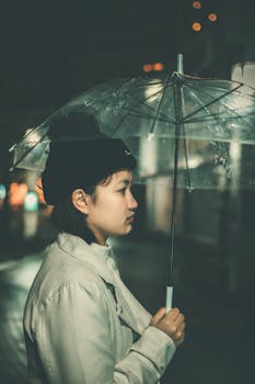 A thoughtful woman stands under a transparent umbrella during a rainy night.