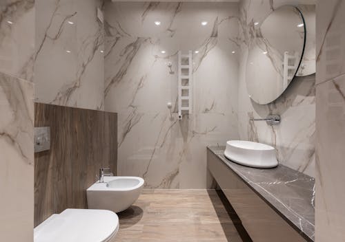 Interior of light modern bathroom with bidet and toilet and sink on cabinet under round mirror on tile