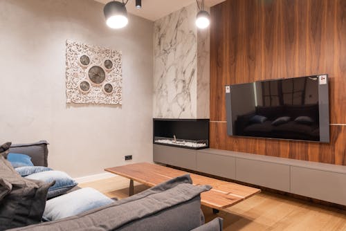 Flat screen TV hanging on wooden wall near fireplace against soft sofa with cushions in modern apartment