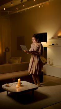 A woman in a dress using a laptop in a warmly lit living room, creating a cozy atmosphere with candles.