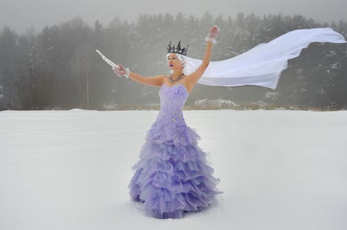 Full body of young graceful female wearing crown and purple dress with icicle in hand and white veil while standing on snowy field near forest in winter day