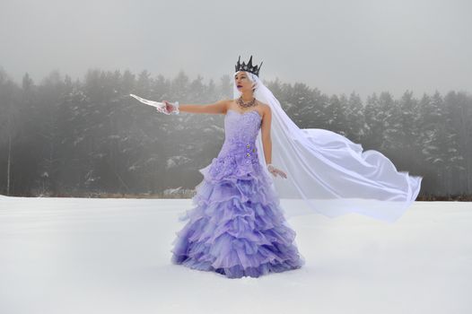 Full length of young elegant female wearing crown and gloves with purple dress and white veil standing on snowy meadow with icicle in hand near woods in winter day