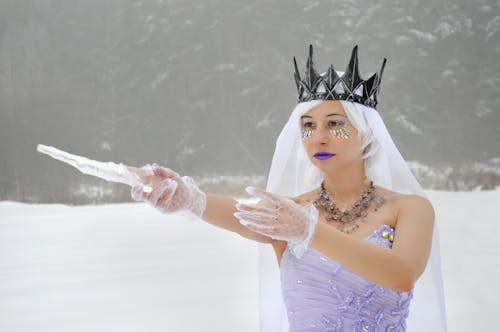 Free Confident young lady in purple dress and white veil with crown with icicle in raised hand in gloves with makeup and crystals on face on snow in winter near forest Stock Photo