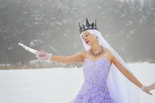Elegant young woman wearing crown and gloves with purple dress and white veil with makeup and crystals on face with icicle in hand on snowy meadow near woods in daytime