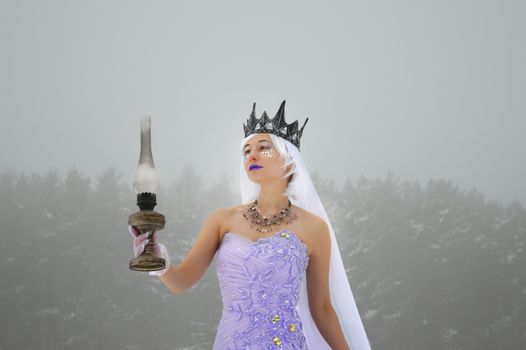 Elegant young woman wearing purple dress and crown with gloves and white veil holding old kerosene lamp in winter snowfall