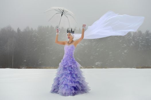 Full length young lady in purple dress and white waving veil with crown and raised hands with transparent umbrella with gloves on snowy field in winter day near forest