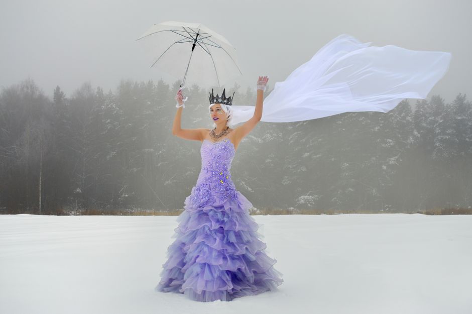 Full length young lady in purple dress and white waving veil with crown and raised hands with transparent umbrella with gloves on snowy field in winter day near forest