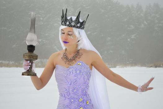 Elegant young woman wearing purple dress and crown with white veil and gloves with old kerosene lamp in hand on snowy meadow near woods