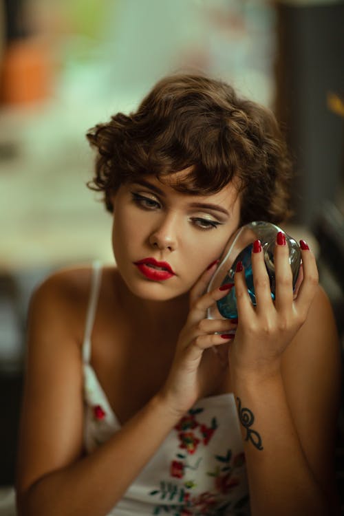 Concentrated female with makeup and short hair wearing dress listening to decorative glass conch while sitting in room on blurred background