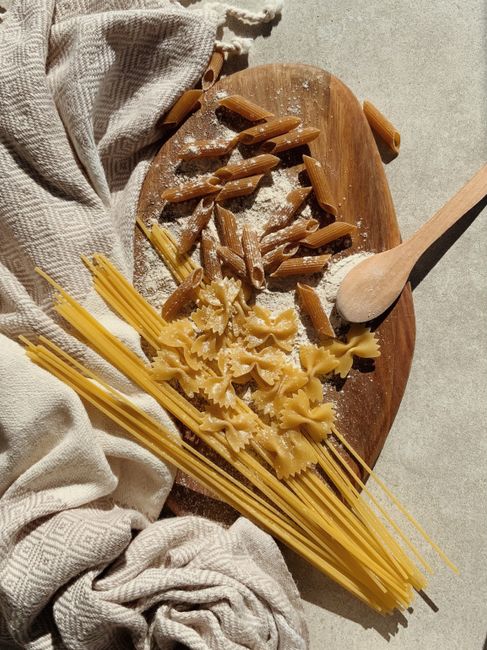 An assortment of dried Italian pasta shapes, including spaghetti, penne, and orecchiette, arranged on a wooden board with a small bowl of semolina flour.