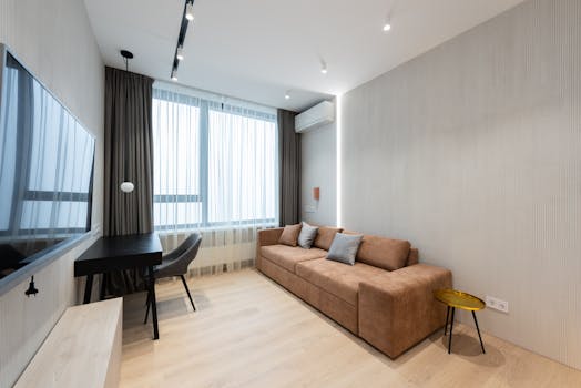 Interior of modern living room with sofa with pillows and TV on wall next to table with chair and curtains on window