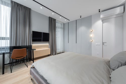 Interior of bright modern apartment with bed next to table with chair and TV on wall near windows with curtains