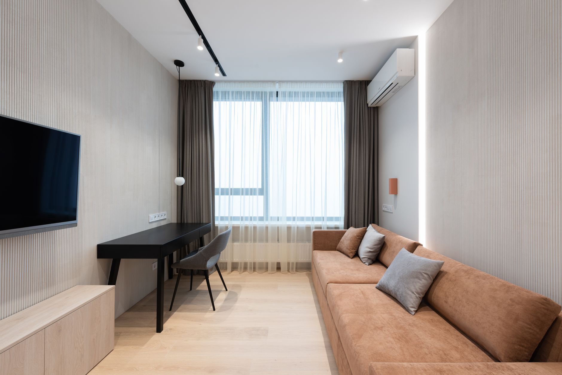 Warm minimalist living room with tan sofa, black desk and chair, wall-mounted TV, and sheer-curtained window.