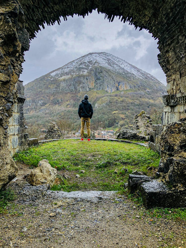 A Back View Of A Person Standing Near The Archway While Facing The Mountain