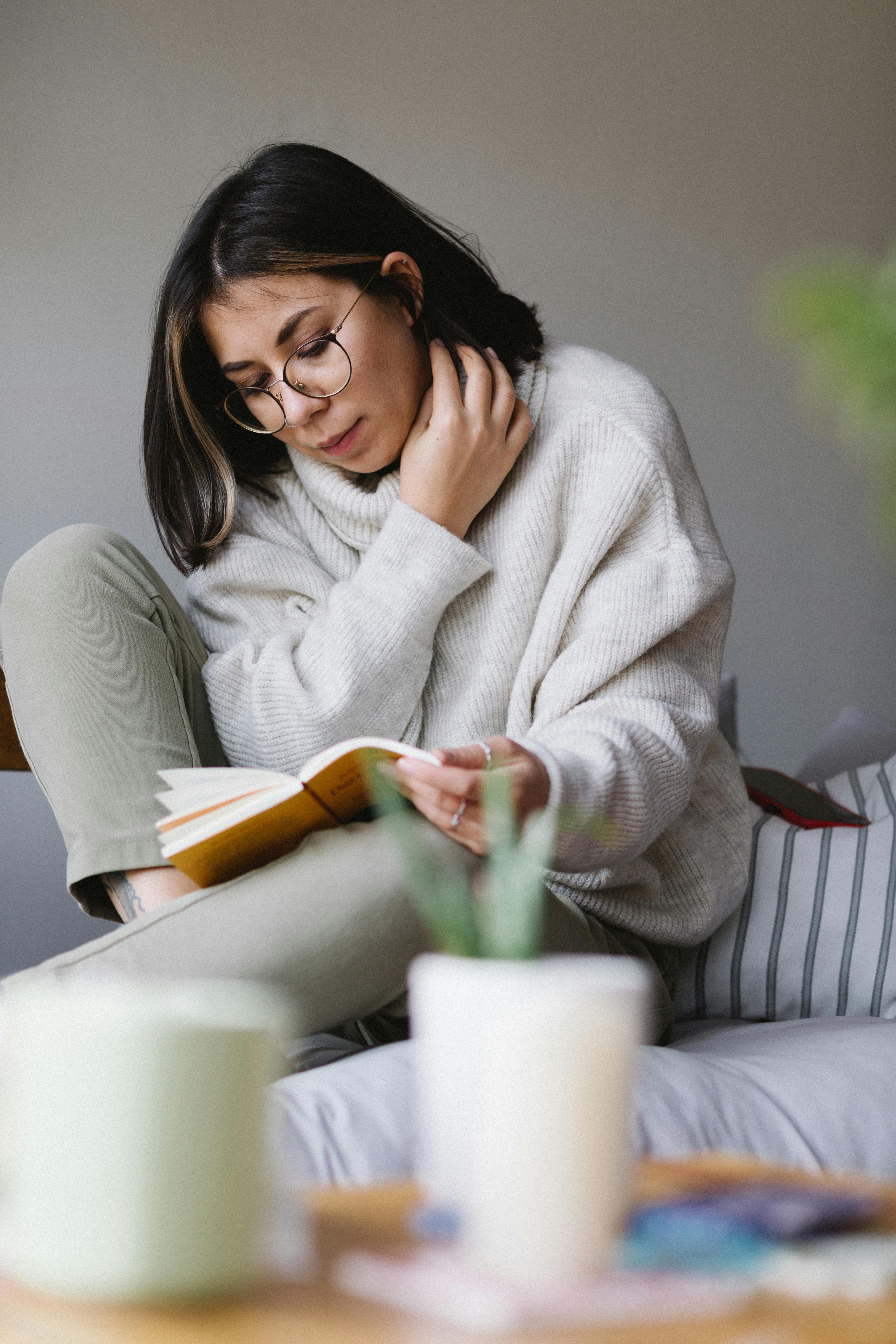Free A young woman in glasses enjoys reading a book on a cozy sofa, epitomizing relaxation and intellect. Stock Photo