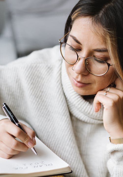 Crop smart female young student in warm casual sweater and eyeglasses writing in notebook while doing homework assignment