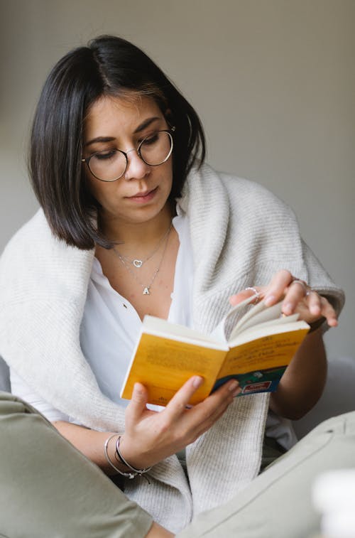 Concentrated young woman with dark hair in casual clothes and eyeglasses turning page of book during reading at home