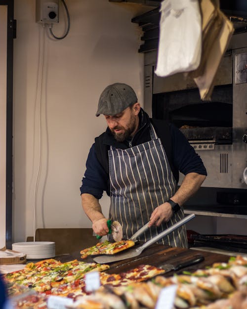 A Bearded Man Wearing Apron while Holding a Pizza and a Cutter