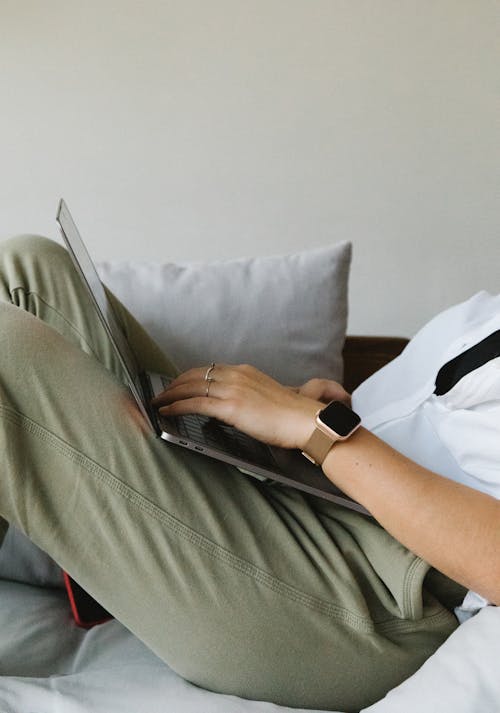 Side view of crop unrecognizable female freelancer in casual clothes and smartwatch lying on comfortable sofa and working remotely on laptop