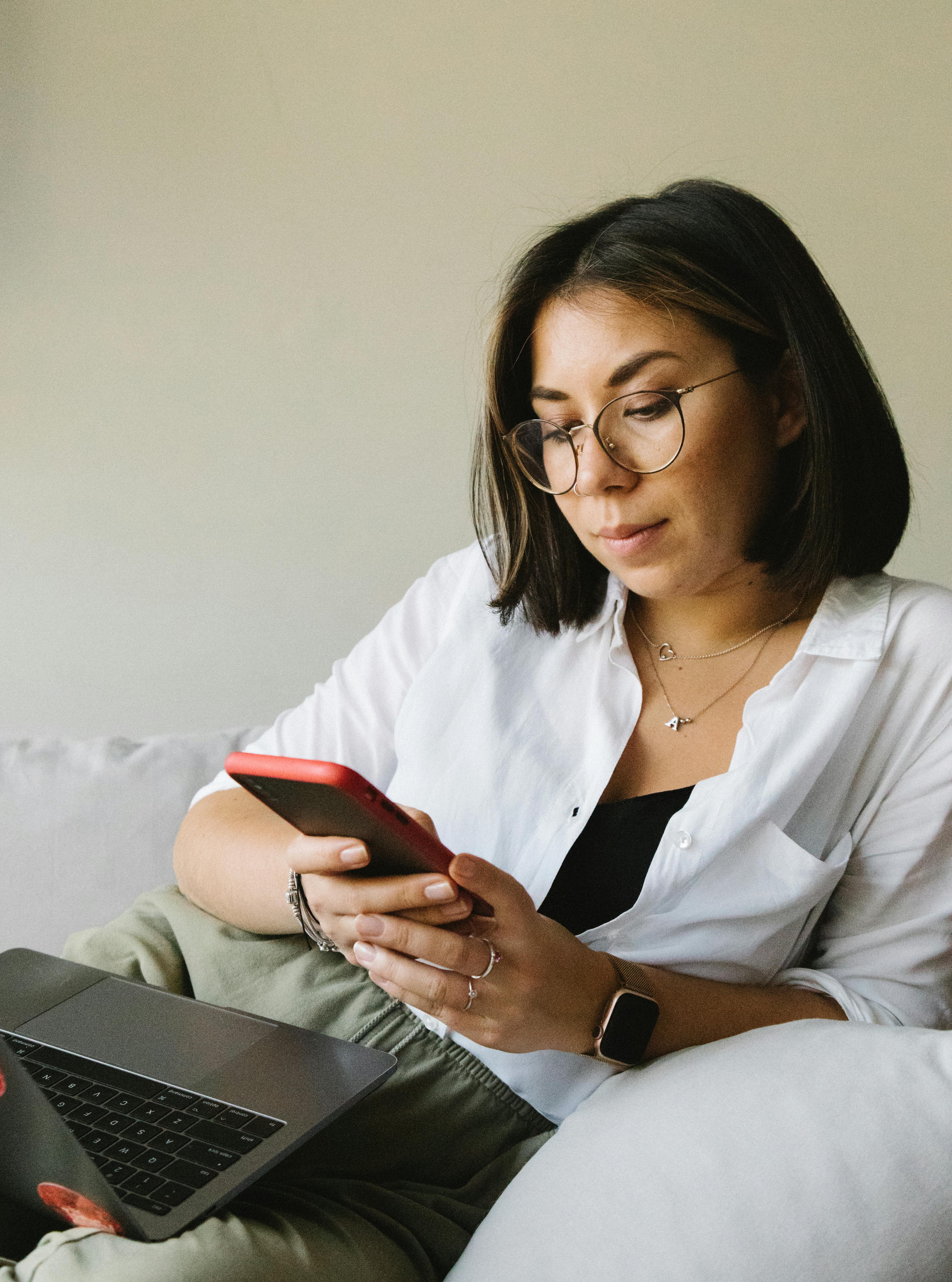 positive woman using smartphone and lying on sofa with laptop