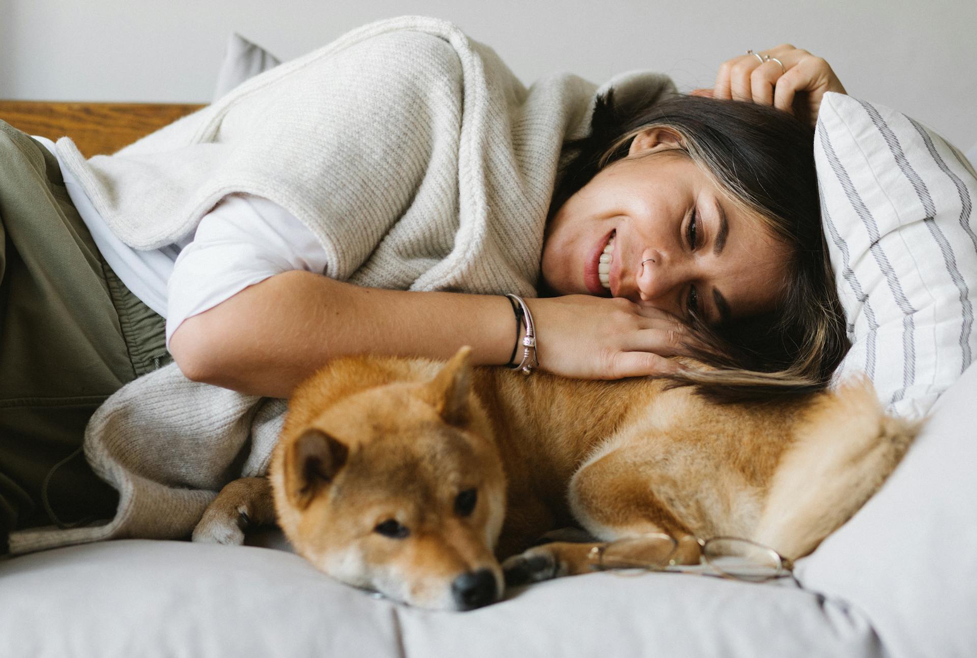 Une brune joyeuse dans des vêtements confortables embrasse un adorable chien Shiba Inu moelleux tout en s'allongeant ensemble sur un canapé confortable dans un salon léger