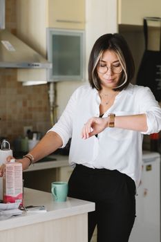 Positive young brunette in casual clothes checking time on wristwatch while standing near counter in modern kitchen