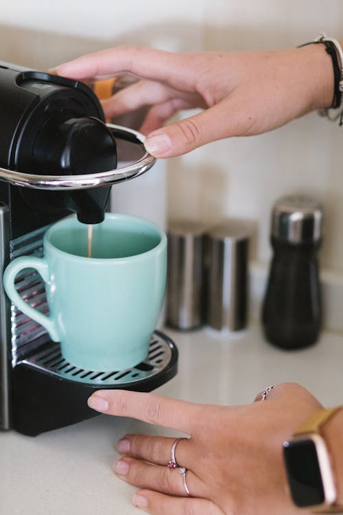 Crop anonymous female brewing hot aromatic coffee by using pod machine in light kitchen