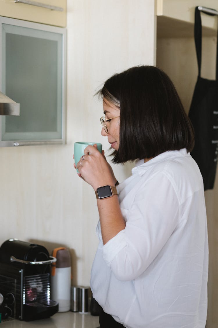 Smiling Young Asian Woman Drinking Coffee From Cup In Kitchen