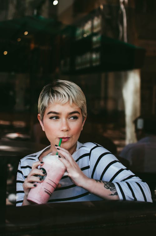 Young glad female with stylish haircut resting with milky cocktail while looking away in cafe