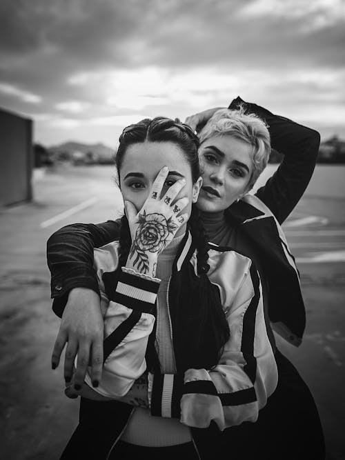 Black and white of young woman hiding face with tattooed hand near friend looking at camera on blurred background