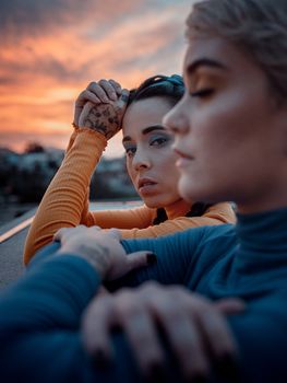 Crop young female closing eyes while friend with tattooed hand looking at camera under shiny cloudy sky