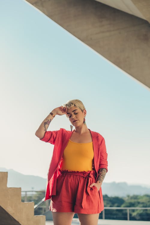 Young female with trendy short hairstyle touching forehead near concrete construction and steps under cloudless shiny sky