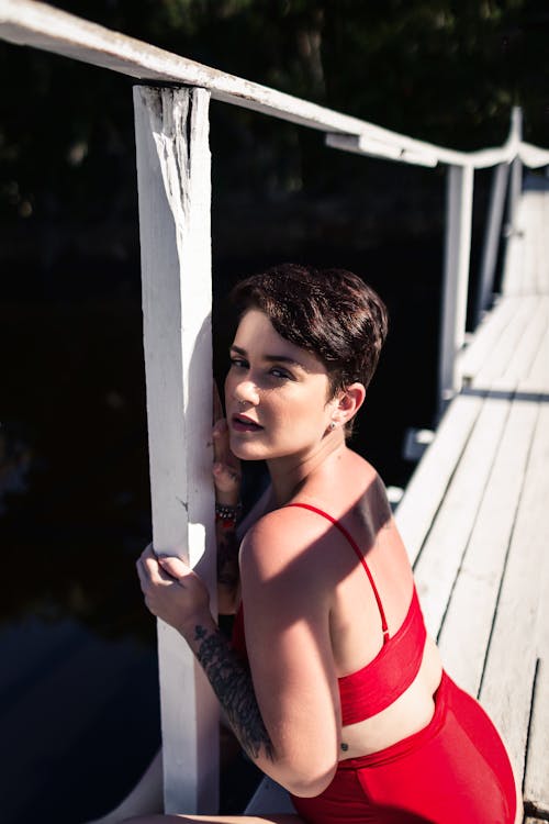 Free Young content female with short dark hair in swimwear looking at camera on timber bridge of river Stock Photo