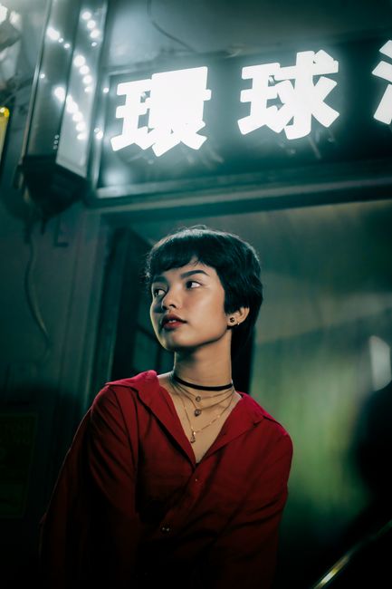 Young ethnic female with short dark hair in trendy red blouse looking away under shiny signboard with hieroglyphs