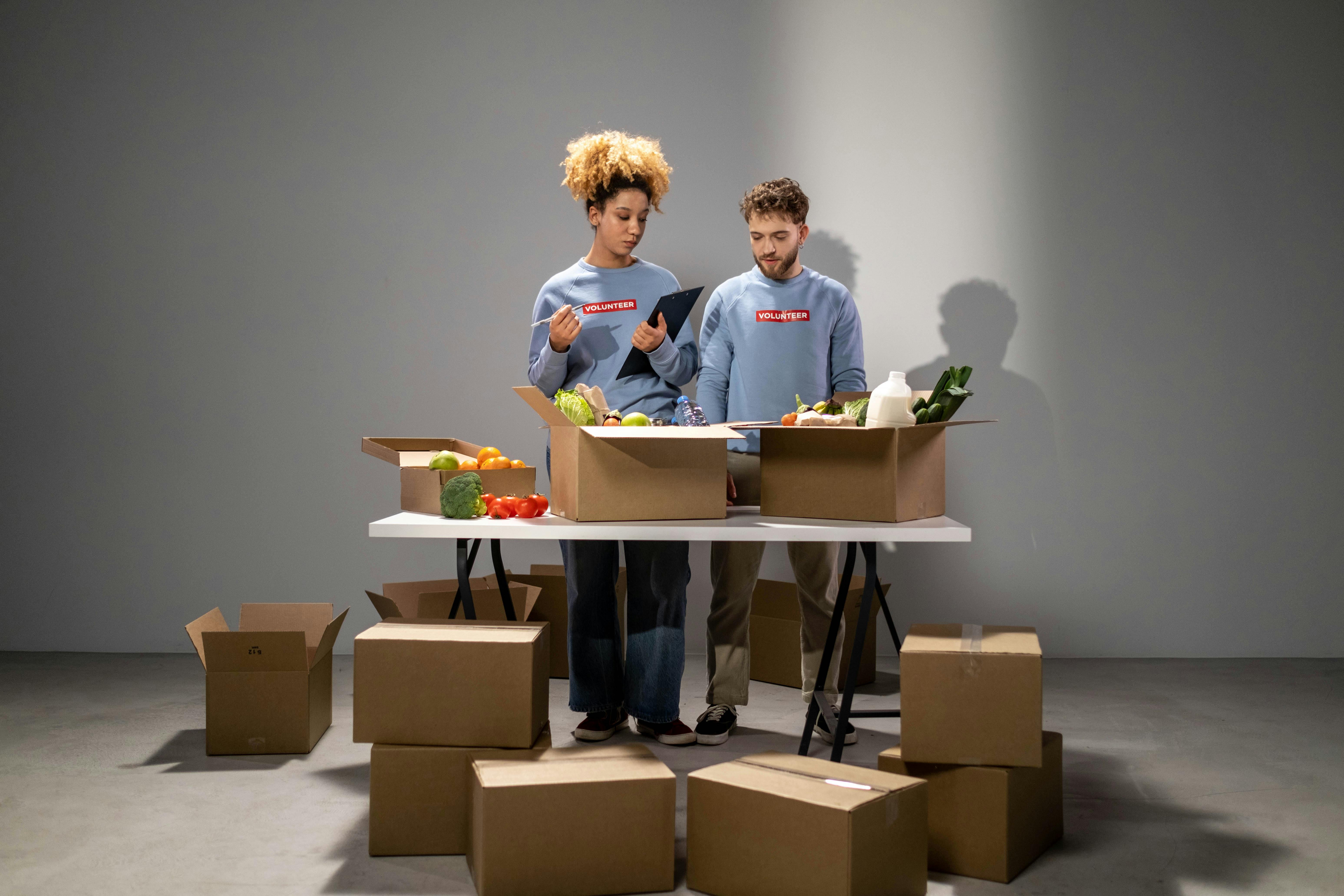 Two volunteers organizing food donations in cardboard boxes for charity indoors.