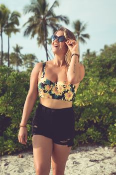 Young glad female in trendy summer outfit and sunglasses on sunny sandy beach with green bushes and palms
