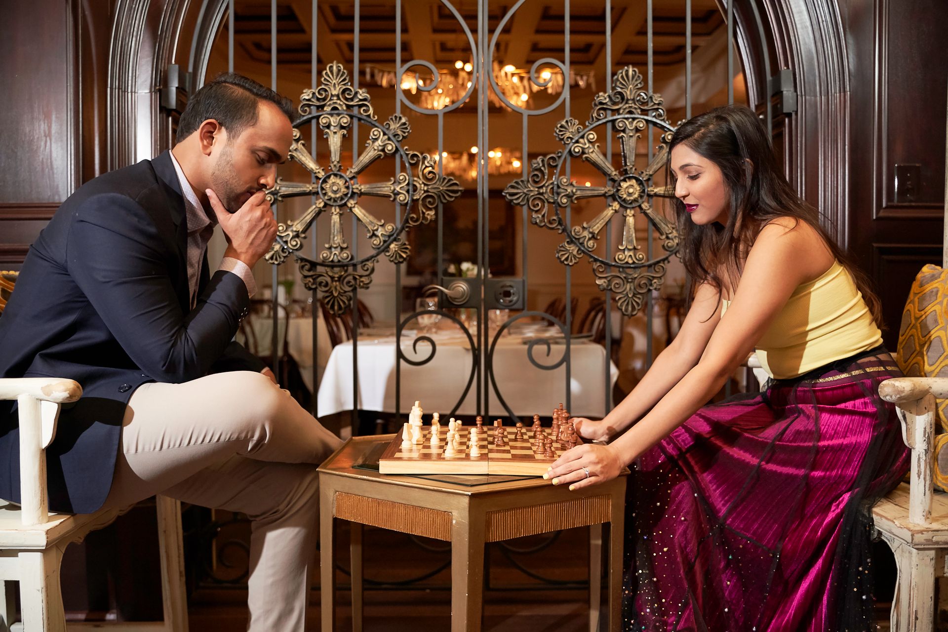 A man and woman engaged in a thoughtful chess game inside a luxurious indoor setting.