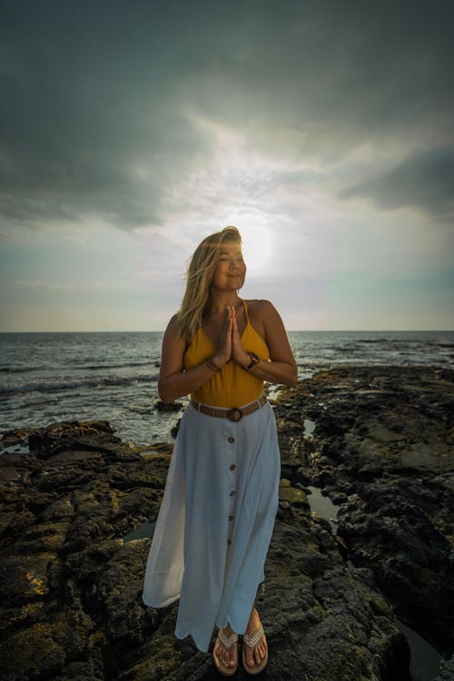 Full body of peaceful female with folded hands doing surya namaskar asana with closed eyes while standing on stony beach near sea