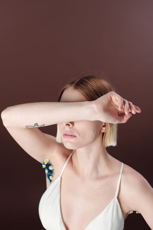Free A young woman poses in a studio with her arm covering her eyes, showcasing a tattoo on a neutral background. Stock Photo