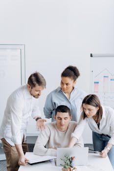 A diverse group of coworkers gathered around a laptop discussing an office project.