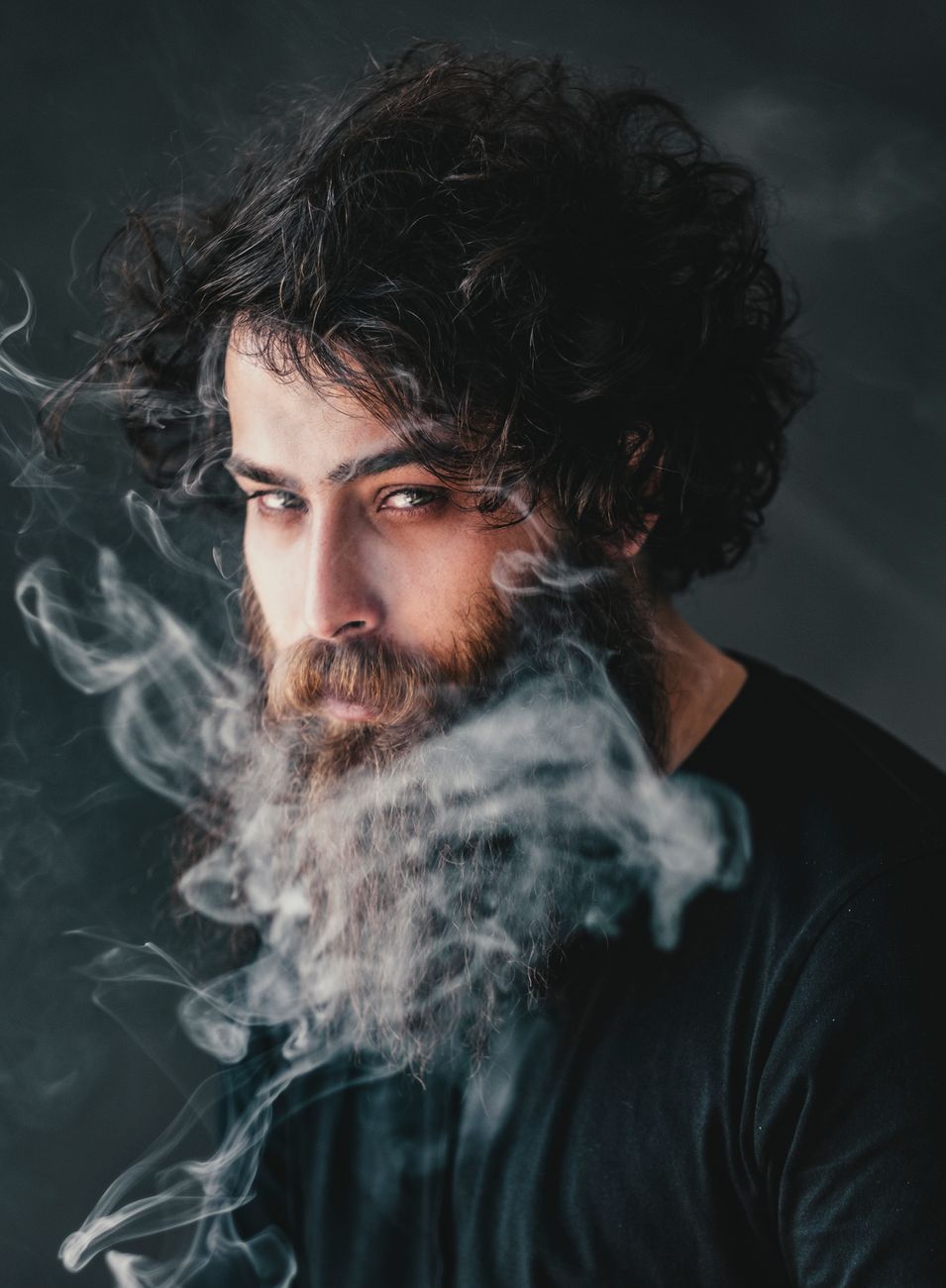 Artistic portrait of a bearded man enveloped in smoke, set against a dark background, capturing a fierce expression.