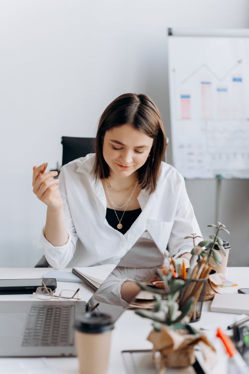 Free A young woman in a white dress shirt working at a desk with a laptop and coffee. Stock Photo
