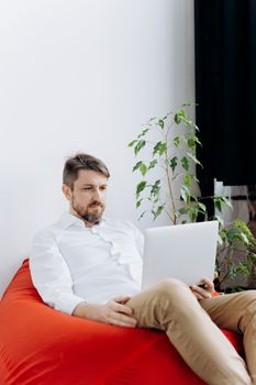 Casual portrait of a man using a laptop while seated on a red bean bag indoors.