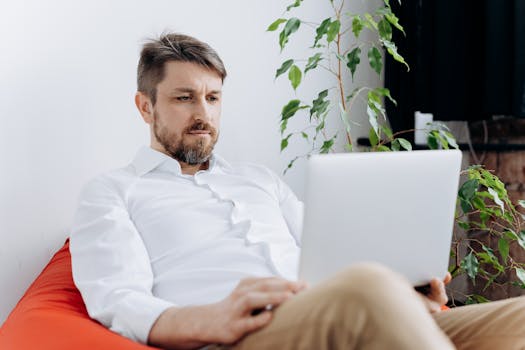 Adult man in a white shirt focused on a laptop while sitting indoors.