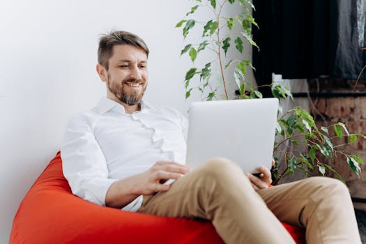 Man sitting on red bean bag chair using laptop, smiling in casual modern workspace.