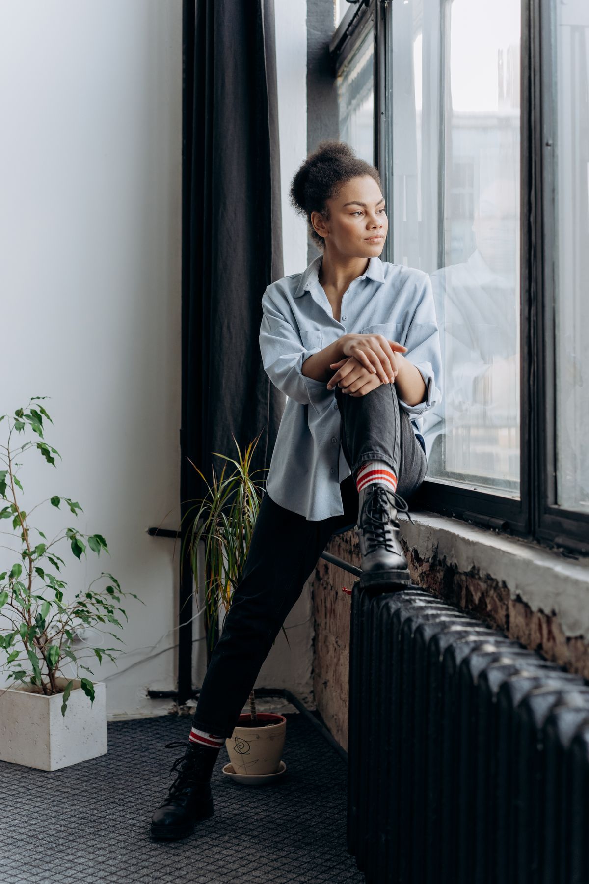 A Black woman with afro hair sits on the windowsill of an older urban apartment, looking out through tall windows in natural afternoon light; a cast-iron radiator and houseplants frame the scene.