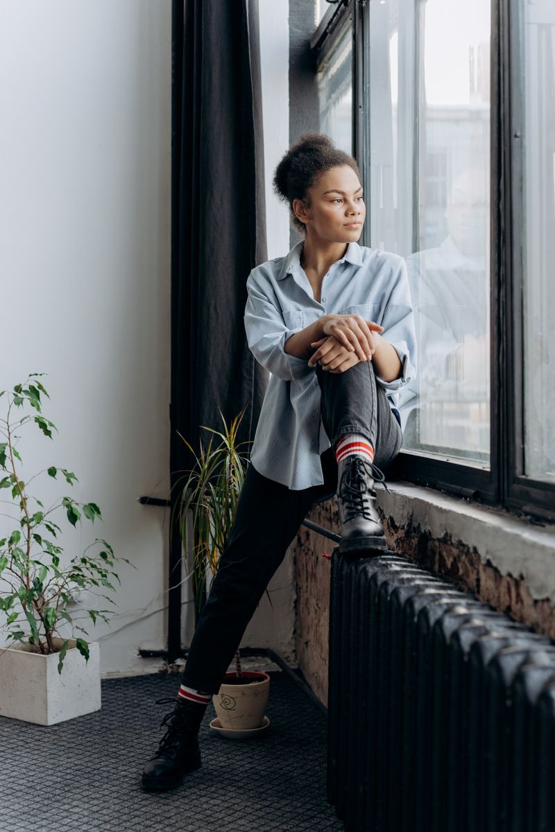 A Black woman with afro hair sits on the windowsill of an older urban apartment, looking out through tall windows in natural afternoon light; a cast-iron radiator and houseplants frame the scene.