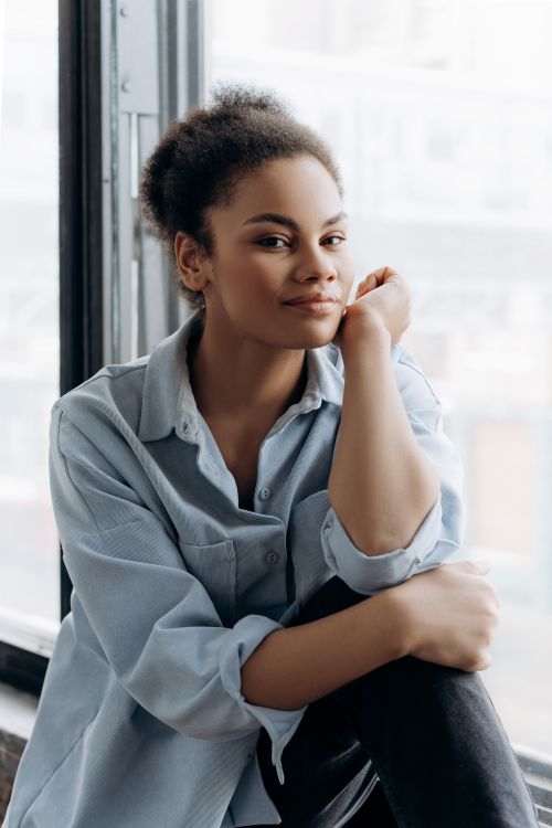 Free Happy young woman sitting by a window wearing a blue shirt, smiling gently. Stock Photo