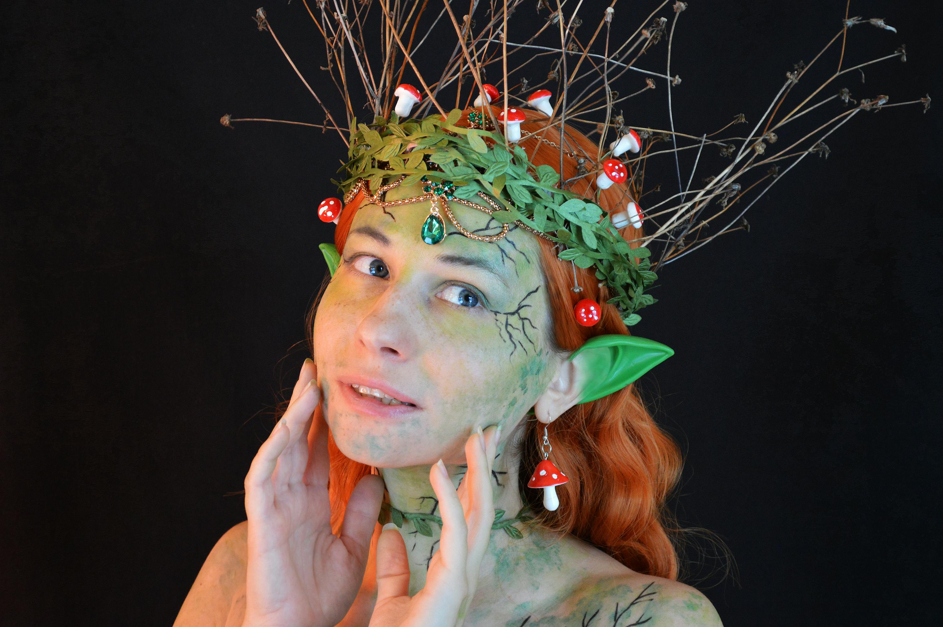 Free Young lady wearing enchantress costume with body art and green wreath with leaves and twigs with small decorative mushrooms in hair and fake ears looking away on black background Stock Photo