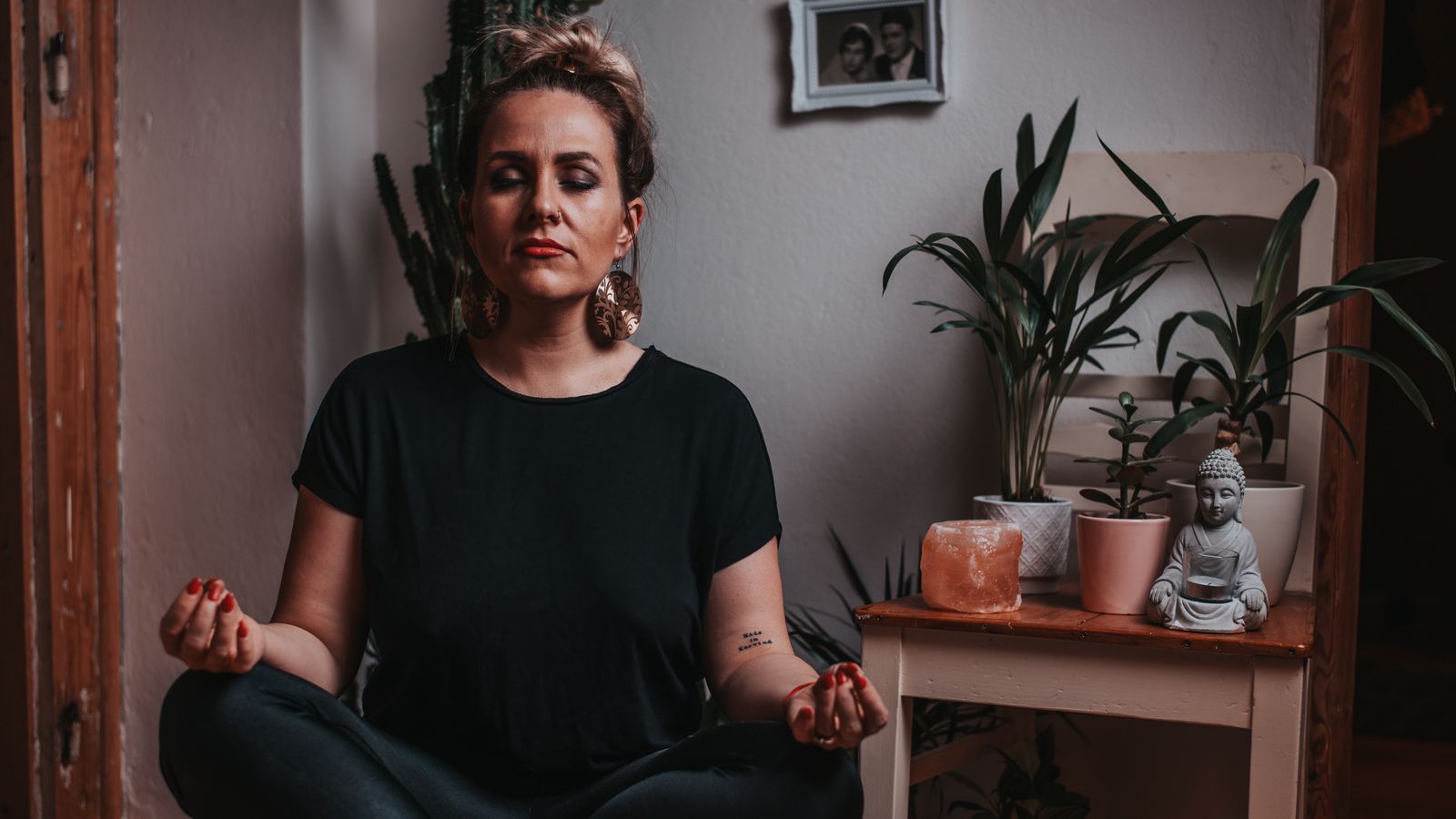 A woman meditating cross-legged indoors with her eyes closed beside houseplants and a Buddha statue